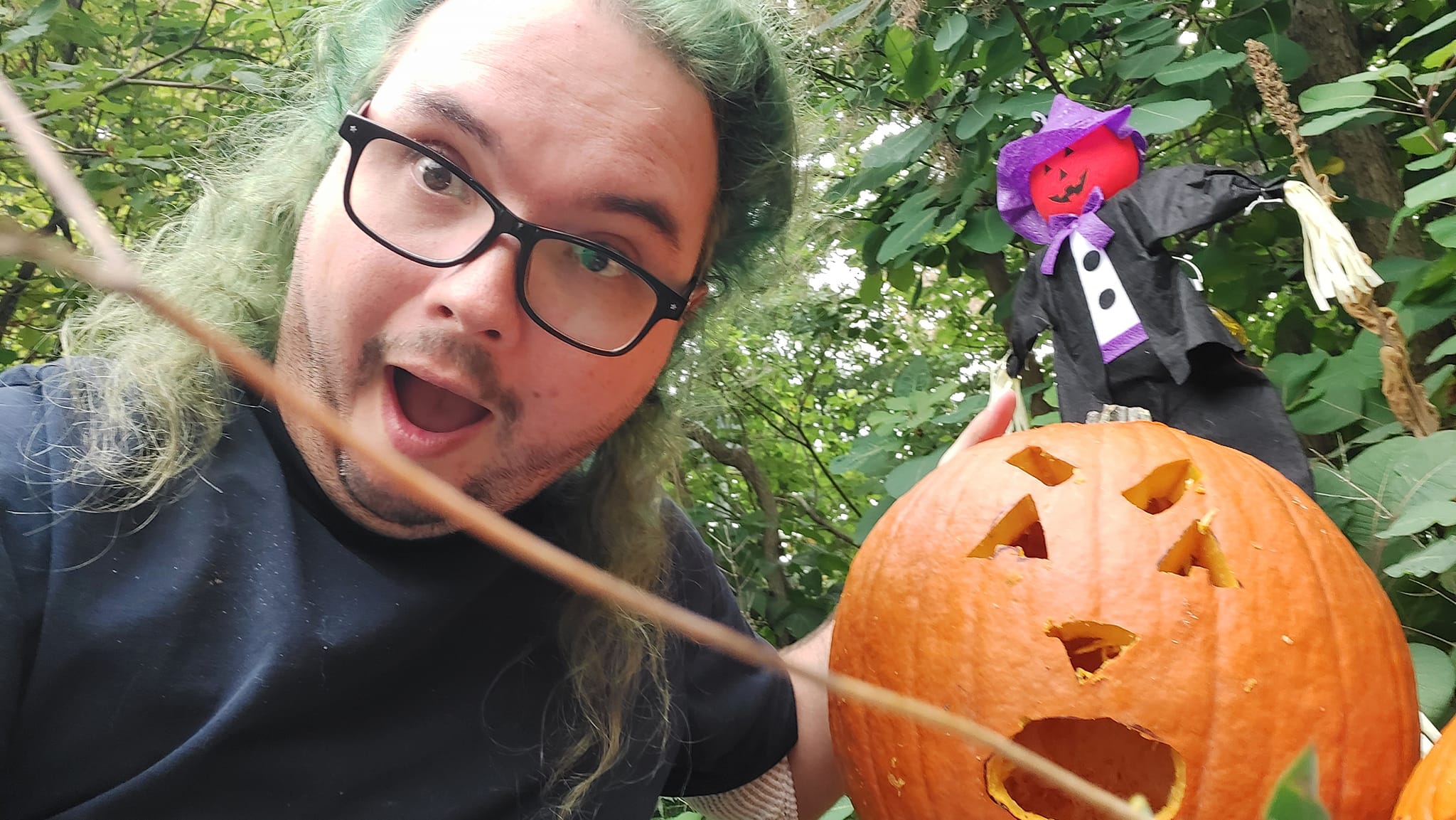 A man with long green hair and no beard holds a large pumpkin with a surprised face carved on it